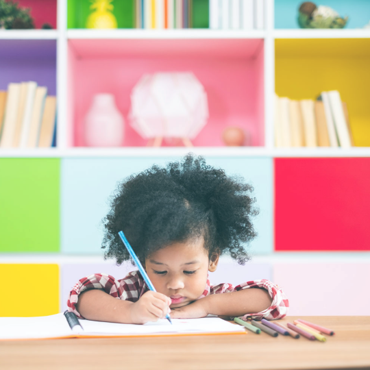Image of young child sitting at table using colored markers on paper in front of colorful bookshelf.